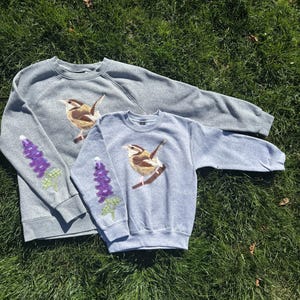 K&ouml;nnte beinhalten: Zwei graue Sweatshirts mit gestickten Designs. Eines ist dunkelgrau, das andere hellblau-grau. Jedes zeigt einen Vogel auf einem Ast und ein lila Blumenmuster auf dem &Auml;rmel. Die Sweatshirts liegen auf gr&uuml;nem Gras.