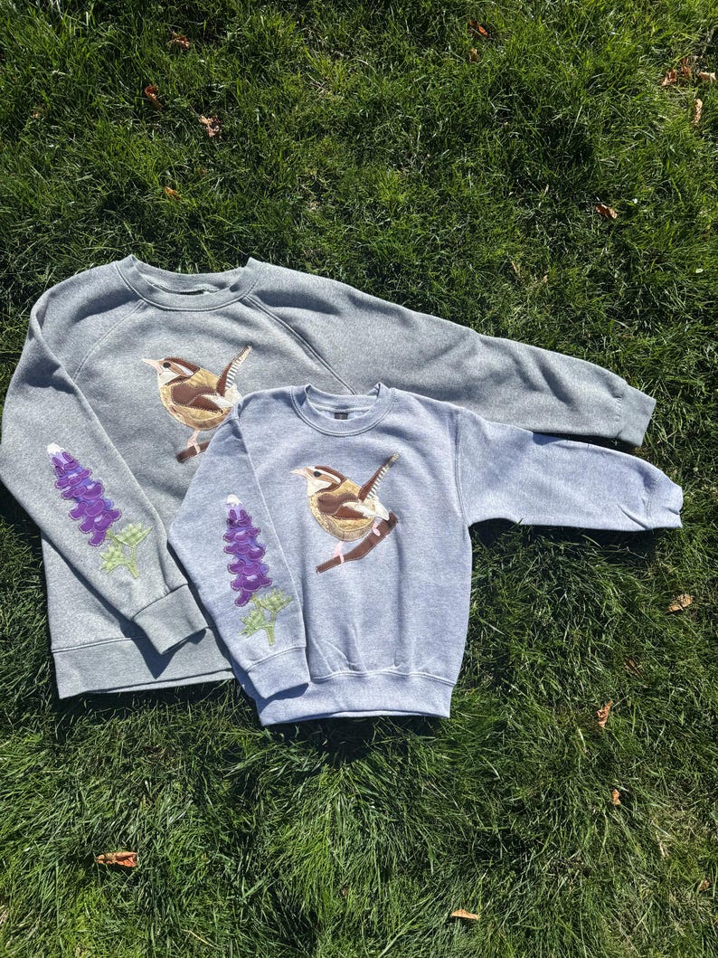 K&ouml;nnte beinhalten: Zwei graue Sweatshirts mit gestickten Designs. Eines ist dunkelgrau, das andere hellblau-grau. Jedes zeigt einen Vogel auf einem Ast und ein lila Blumenmuster auf dem &Auml;rmel. Die Sweatshirts liegen auf gr&uuml;nem Gras.
