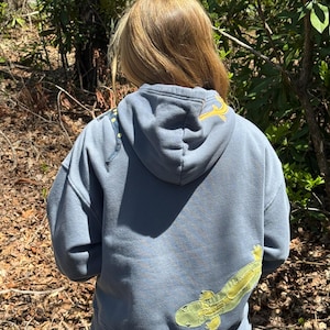 K&ouml;nnte beinhalten: Ein blau-grauer Kapuzenpullover mit einem gro&szlig;en, hellgr&uuml;nen und gelben Salamander-Applikation auf dem R&uuml;cken. Die Kapuze hat ein gelbes Stickdesign. Der Pullover wird im Freien in einem Waldgebiet getragen.