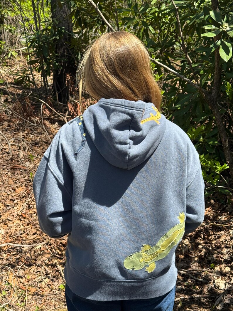 K&ouml;nnte beinhalten: Ein blau-grauer Kapuzenpullover mit einem gro&szlig;en, hellgr&uuml;nen und gelben Salamander-Applikation auf dem R&uuml;cken. Die Kapuze hat ein gelbes Stickdesign. Der Pullover wird im Freien in einem Waldgebiet getragen.