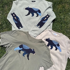 K&ouml;nnte beinhalten: Vier Kleidungsst&uuml;cke: zwei Sweatshirts und zwei T-Shirts. Jedes Kleidungsst&uuml;ck hat eine dunkelblaue B&auml;renapplikation mit hellblauen Akzenten. Die Sweatshirts sind hellgr&uuml;n, und die T-Shirts sind oliv und beige.