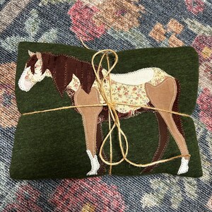 K&ouml;nnte beinhalten: Gefalteter gr&uuml;ner Stoff mit einem Pferde-Applikation. Das Pferdedesign weist braune, beige und floral gemusterte Stoffdetails auf. Das B&uuml;ndel ist mit Bindfaden gebunden. Der Hintergrund ist ein Stoff mit floralem Muster.
