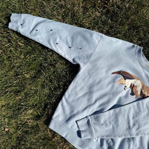 K&ouml;nnte beinhalten: Hellblaues Sweatshirt mit gesticktem Ameisenb&auml;ren-Design auf der Vorderseite. Das Sweatshirt hat lange &Auml;rmel mit gestickten schwarzen Insekten auf dem linken &Auml;rmel. Das Kleidungsst&uuml;ck ist aus weichem Material.