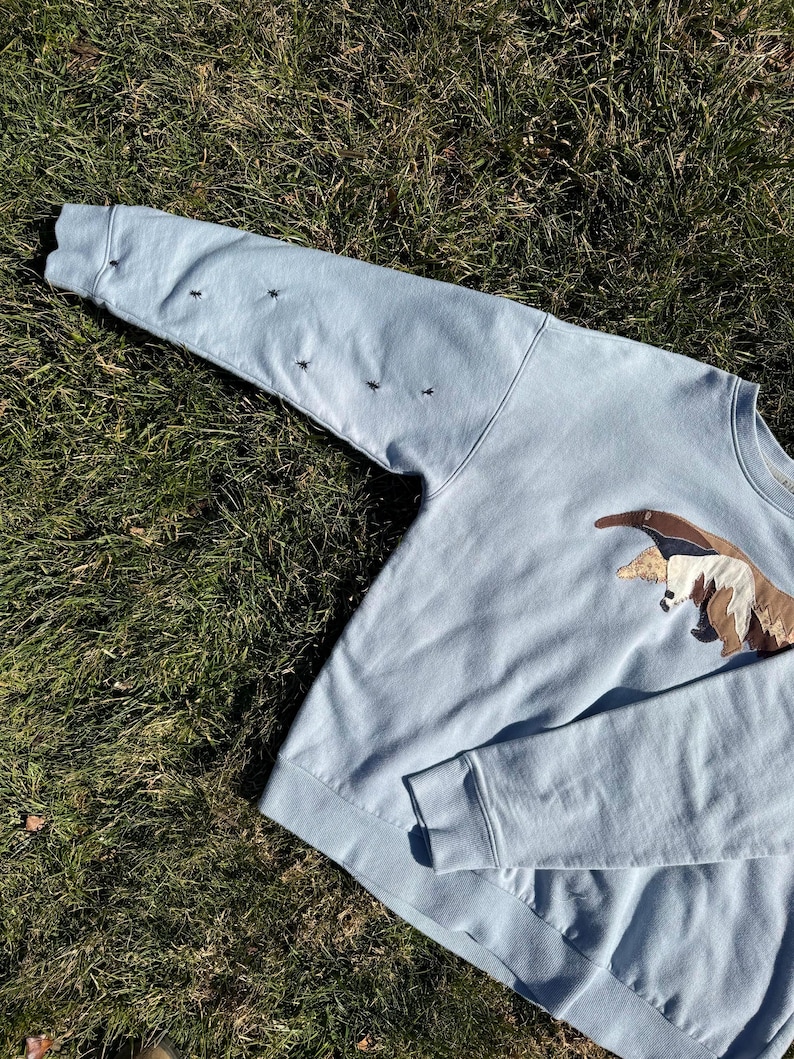 K&ouml;nnte beinhalten: Hellblaues Sweatshirt mit gesticktem Ameisenb&auml;ren-Design auf der Vorderseite. Das Sweatshirt hat lange &Auml;rmel mit gestickten schwarzen Insekten auf dem linken &Auml;rmel. Das Kleidungsst&uuml;ck ist aus weichem Material.