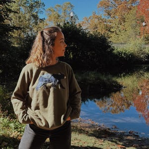 K&ouml;nnte beinhalten: Ein olivgr&uuml;nes Sweatshirt mit einem dunkelblauen Hundeaufn&auml;her. Die Person tr&auml;gt eine schwarze Jeans und steht drau&szlig;en in der N&auml;he eines Gew&auml;ssers. Der Hintergrund umfasst B&auml;ume mit Herbstlaub und einen klaren blauen Himmel.
