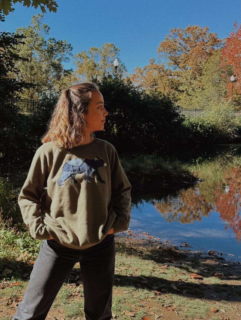 K&ouml;nnte beinhalten: Ein olivgr&uuml;nes Sweatshirt mit einem dunkelblauen Hundeaufn&auml;her. Die Person tr&auml;gt eine schwarze Jeans und steht drau&szlig;en in der N&auml;he eines Gew&auml;ssers. Der Hintergrund umfasst B&auml;ume mit Herbstlaub und einen klaren blauen Himmel.