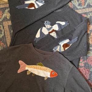 K&ouml;nnte beinhalten: Dunkelgraue Sweatshirts mit Applikationsdesigns. Ein Sweatshirt zeigt einen Fisch mit rot-wei&szlig; kariertem Schwanz. Die anderen Sweatshirts haben Vogelmotive mit blauen, wei&szlig;en und braunen Akzenten. Die Sweatshirts sind auf einem gemusterten Teppich gefaltet.