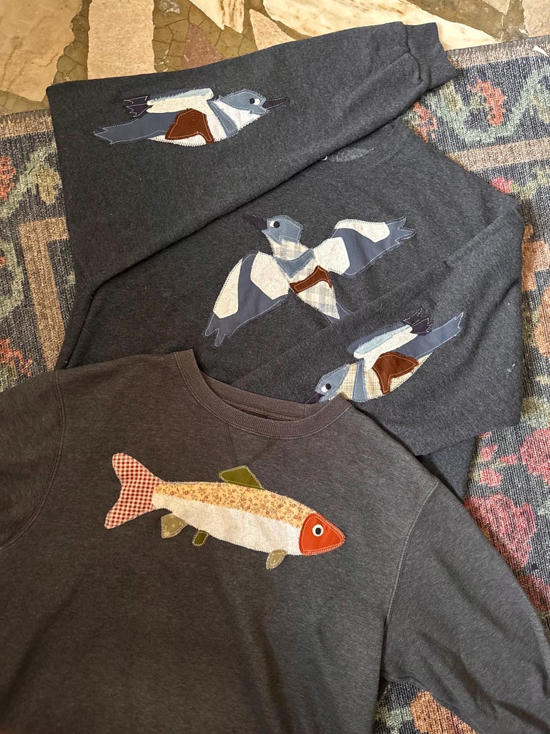 K&ouml;nnte beinhalten: Dunkelgraue Sweatshirts mit Applikationsdesigns. Ein Sweatshirt zeigt einen Fisch mit rot-wei&szlig; kariertem Schwanz. Die anderen Sweatshirts haben Vogelmotive mit blauen, wei&szlig;en und braunen Akzenten. Die Sweatshirts sind auf einem gemusterten Teppich gefaltet.