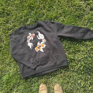 K&ouml;nnte beinhalten: Schwarzes Sweatshirt mit Rundhalsausschnitt und einem kreisf&ouml;rmigen Design aus zwei Koi-Karpfen in Rot, Wei&szlig; und Orange. Das Sweatshirt ist aus weichem Material und liegt auf gr&uuml;nem Gras.