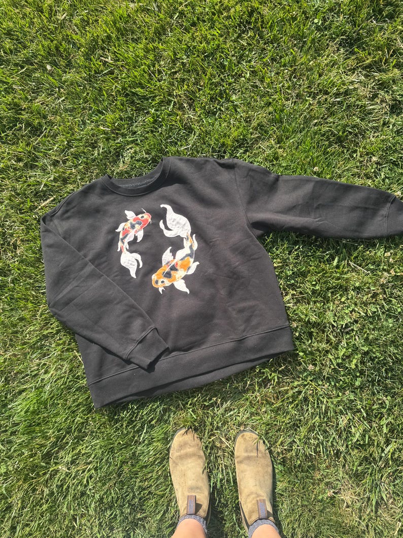 K&ouml;nnte beinhalten: Schwarzes Sweatshirt mit Rundhalsausschnitt und einem kreisf&ouml;rmigen Design aus zwei Koi-Karpfen in Rot, Wei&szlig; und Orange. Das Sweatshirt ist aus weichem Material und liegt auf gr&uuml;nem Gras.