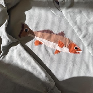 K&ouml;nnte beinhalten: Hellblaues Sweatshirt mit aufgen&auml;htem Fisch-Applikation. Das Fischdesign weist Braunt&ouml;ne, Orange und Wei&szlig; auf, mit detailliertem Auge und Flossen. Das Sweatshirt hat einen Rundhalsausschnitt und lange &Auml;rmel.