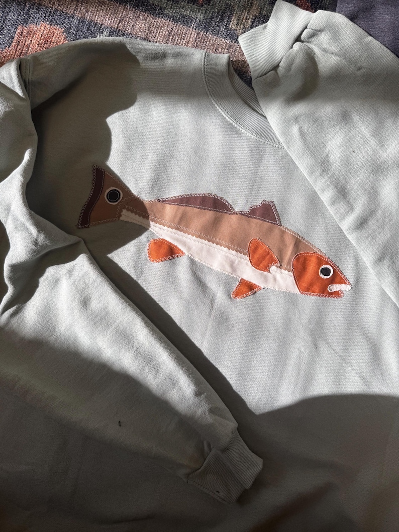 K&ouml;nnte beinhalten: Hellblaues Sweatshirt mit aufgen&auml;htem Fisch-Applikation. Das Fischdesign weist Braunt&ouml;ne, Orange und Wei&szlig; auf, mit detailliertem Auge und Flossen. Das Sweatshirt hat einen Rundhalsausschnitt und lange &Auml;rmel.