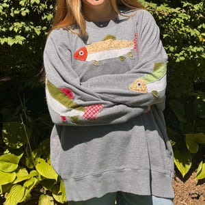 K&ouml;nnte beinhalten: Ein graues Sweatshirt mit aufgen&auml;hten Fischmotiven in verschiedenen Farben. Die Fische haben beige, gr&uuml;ne, rote und wei&szlig;e Akzente. Das Sweatshirt ist ein l&auml;ssiger Stil, der f&uuml;r den Alltag geeignet ist.