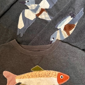 K&ouml;nnte beinhalten: Dunkelgraue Sweatshirts mit Applikationsdesigns. Ein Sweatshirt zeigt einen Fisch mit rot-wei&szlig; kariertem Schwanz und einem floralen K&ouml;rper. Die anderen Sweatshirts haben applizierte V&ouml;gel mit blau, wei&szlig; und braun gemusterten Fl&uuml;geln.