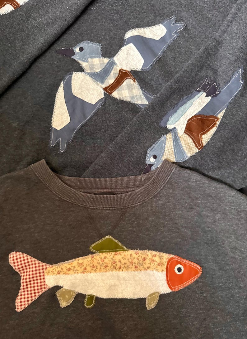 K&ouml;nnte beinhalten: Dunkelgraue Sweatshirts mit Applikationsdesigns. Ein Sweatshirt zeigt einen Fisch mit rot-wei&szlig; kariertem Schwanz und einem floralen K&ouml;rper. Die anderen Sweatshirts haben applizierte V&ouml;gel mit blau, wei&szlig; und braun gemusterten Fl&uuml;geln.