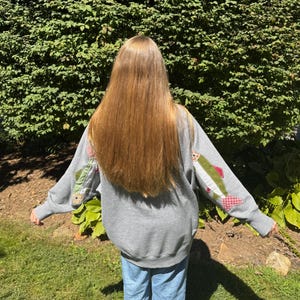 K&ouml;nnte beinhalten: Ein graues Sweatshirt mit fischf&ouml;rmigen Aufn&auml;hern auf den &Auml;rmeln. Die Aufn&auml;her zeigen gr&uuml;nen, roten und wei&szlig;en Stoff. Die Person tr&auml;gt eine blaue Jeans und steht vor einem gr&uuml;nen Busch.