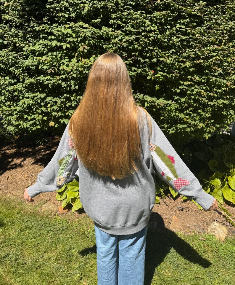 K&ouml;nnte beinhalten: Ein graues Sweatshirt mit fischf&ouml;rmigen Aufn&auml;hern auf den &Auml;rmeln. Die Aufn&auml;her zeigen gr&uuml;nen, roten und wei&szlig;en Stoff. Die Person tr&auml;gt eine blaue Jeans und steht vor einem gr&uuml;nen Busch.