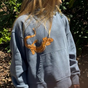 K&ouml;nnte beinhalten: Ein blauer Hoodie mit K&auml;ngurutasche und einem gro&szlig;en, orangefarbenen, gestickten Design einer Meereskreatur auf der Vorderseite. Der Hoodie ist aus einem weichen Material gefertigt und hat eine Kapuze mit Kordelzug.