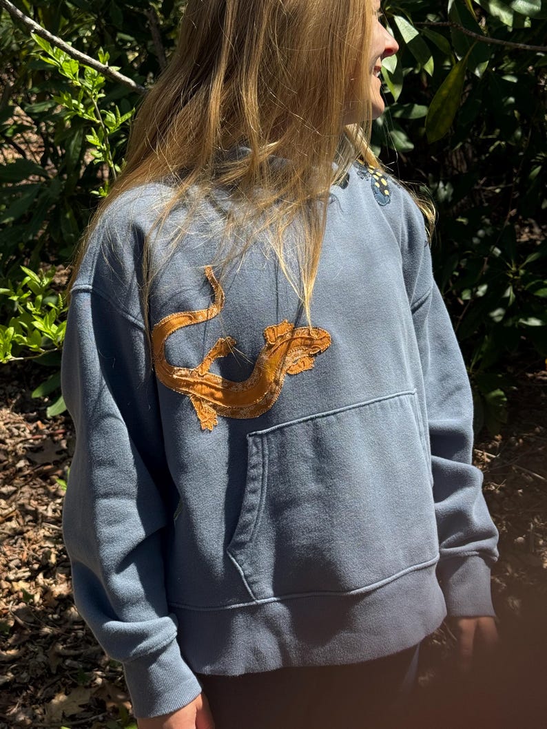 K&ouml;nnte beinhalten: Ein blauer Hoodie mit K&auml;ngurutasche und einem gro&szlig;en, orangefarbenen, gestickten Design einer Meereskreatur auf der Vorderseite. Der Hoodie ist aus einem weichen Material gefertigt und hat eine Kapuze mit Kordelzug.