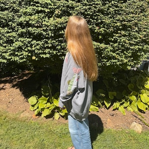 K&ouml;nnte beinhalten: Ein graues Sweatshirt mit einem Fisch-Applikation auf dem R&uuml;cken. Der Fisch hat einen rot-wei&szlig; gef&auml;rbten Schwanz und einen gr&uuml;n-wei&szlig;en K&ouml;rper. Die Person tr&auml;gt eine blaue Jeans und steht vor einem gr&uuml;nen Busch.