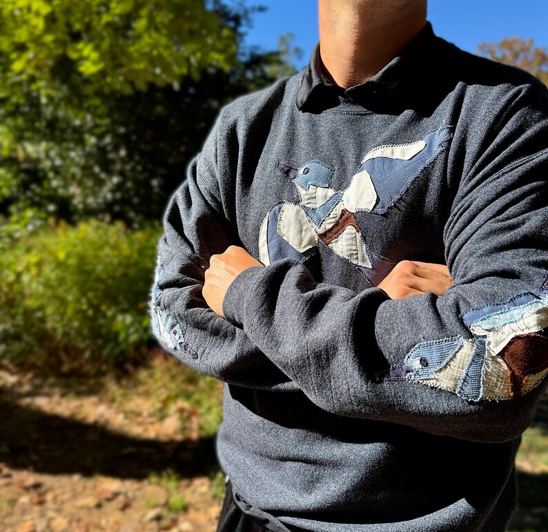 K&ouml;nnte beinhalten: Ein dunkelgraues Sweatshirt mit einem Vogel-Applikation auf der Vorderseite und den &Auml;rmeln. Das Vogeldesign enth&auml;lt blaue, wei&szlig;e und braune Stoffflicken. Die &Auml;rmel haben zus&auml;tzliche Vogelakzente.