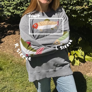 K&ouml;nnte beinhalten: Graues Sweatshirt mit Fischapplikationen auf der Vorderseite und den &Auml;rmeln. Die Vorderseite zeigt ein detailliertes Fischdesign mit roten, beigen und gr&uuml;nen Akzenten. &Auml;rmel mit Fisch- und Karopatch. Ein l&auml;ssiges, bequemes Kleidungsst&uuml;ck.