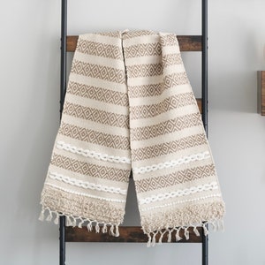 May include: A cream-colored throw blanket with a geometric pattern in brown. The blanket has a fringe border and is hanging on a black metal ladder.