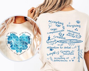 Può includere: Una maglietta color crema con testo e illustrazioni blu. Il testo include "Accepting", "Forgiving", "Compassionate", "Creative", "Honest", "Hyper-focused", "Attention to detail", "Loyal", "Highly Sensitive", "Accepting of differences" e "Independent". La maglietta presenta anche un pezzo di puzzle a forma di cuore blu.