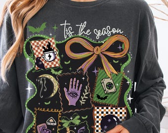 Może przedstawiać: Ciemnoszara bluza z kolorowym wzorem graficznym przedstawiającym kokardę, nietoperze i różne mistyczne symbole. Tekst "Tis the season" jest napisany na górze. Projekt zawiera wzór szachownicy i fioletową dłoń z okiem.