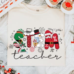 Op de afbeelding: Crèmekleurig T-shirt met een kleurrijk STEM-leraarontwerp. Het ontwerp toont de letters S, T, E en M, elk versierd met vakantie-elementen zoals een elfenmuts, een sneeuwpop en een kerstmuts. Het woord "teacher" staat eronder in cursief.