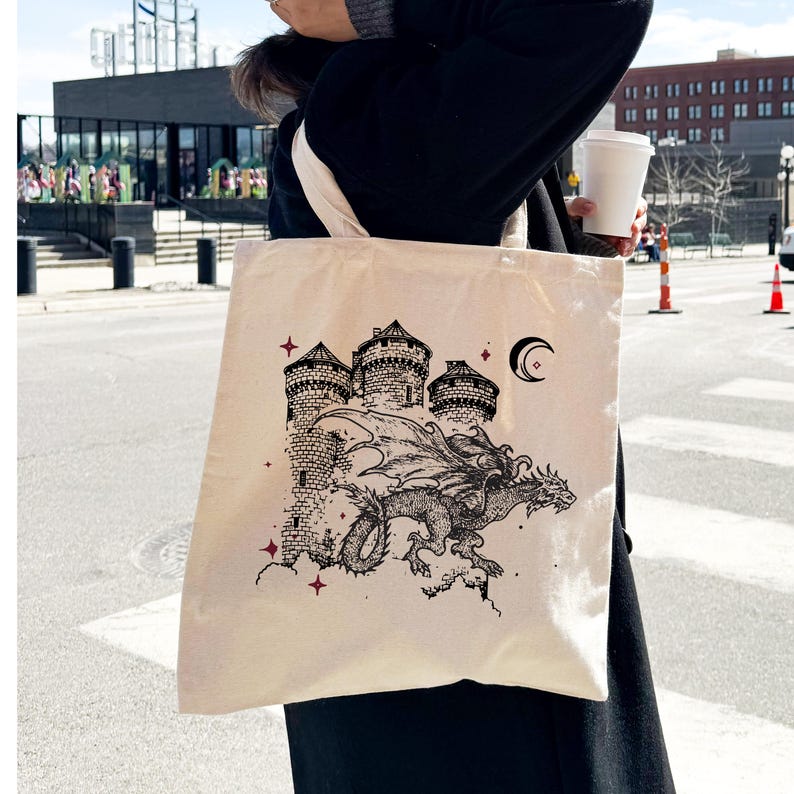 May include: A cream-colored tote bag with a black and white illustration of a dragon flying over a castle. The castle has three towers and a crescent moon is in the sky above the dragon.