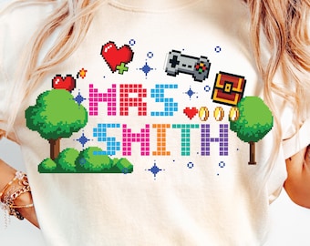 Puede incluir: Una camiseta color crema con un diseño pixelado. El diseño presenta el texto "MRS SMITH" en letras de bloque coloridas, junto con corazones, un mando de juego, un cofre del tesoro, árboles y estrellas, con temática de videojuegos.