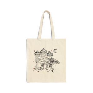 May include: A cream-colored tote bag with a black and white illustration of a dragon flying over a castle with a crescent moon in the sky. The illustration is surrounded by small stars.