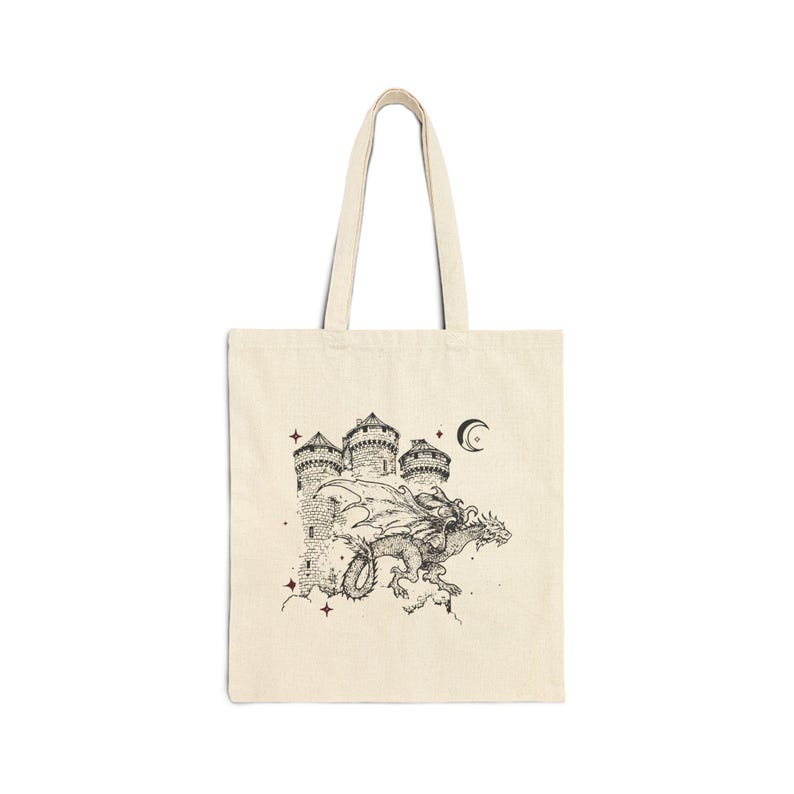 May include: A cream-colored tote bag with a black and white illustration of a dragon flying over a castle with a crescent moon in the sky. The illustration is surrounded by small stars.