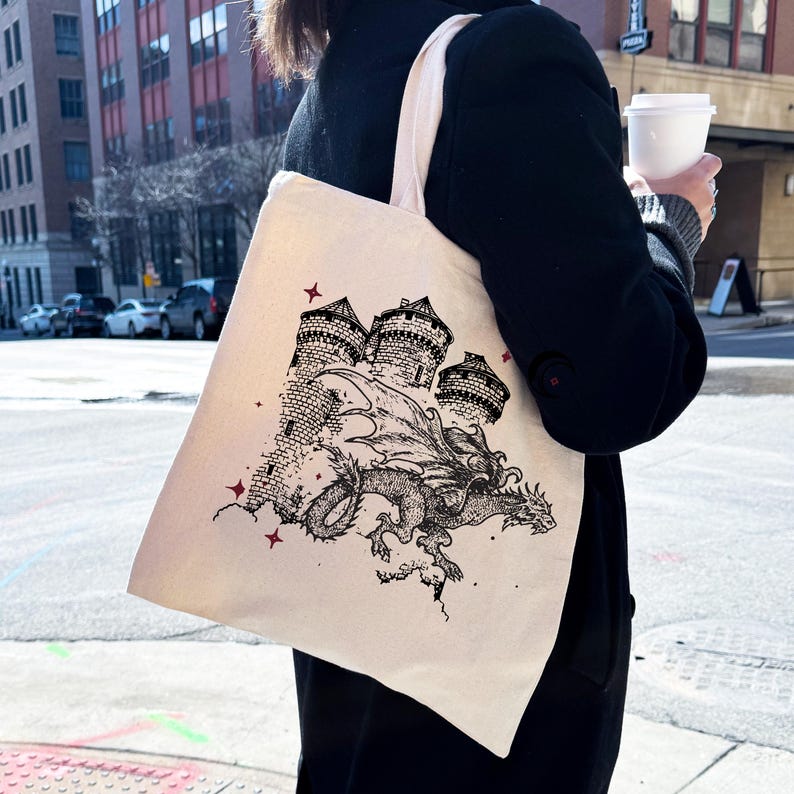May include: A white canvas tote bag with a black and white illustration of a dragon flying over a castle. The dragon has wings and a long tail. The castle has three towers and is surrounded by clouds. There are red stars on the bag.