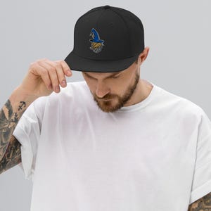 May include: Black baseball cap with a flat brim. The cap features an embroidered design of a wizard hat in blue and gold. The hat is worn by a person in a white t-shirt.