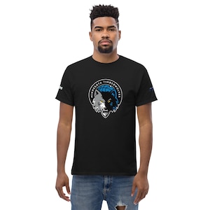 May include: Black t-shirt featuring the Minnesota Timberwolves logo. The logo depicts a wolf's head in white and black, set against a blue basketball and the words "Minnesota Timberwolves" in a circular design.