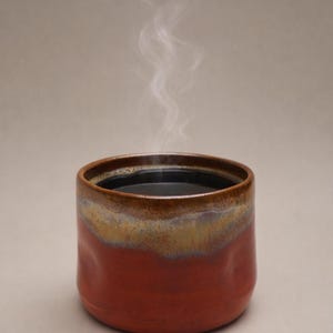 Peut inclure: Une tasse en céramique rouge-orangé avec un bord marron, contenant un liquide sombre. De la vapeur s'élève de la tasse. La tasse a une forme cylindrique et une finition lisse et glacée. Le fond est beige neutre.