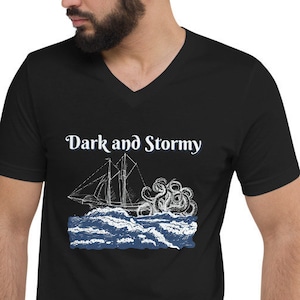 Può includere: T-shirt nero a collo a V con una grafica bianca di una nave che naviga in un mare tempestoso con il testo "Dark and Stormy" sopra l'immagine.