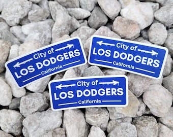May include: Three blue and white rectangular stickers with the text "City of LOS DODGERS California" are displayed on a bed of gray rocks. The stickers feature a road sign design with arrows.
