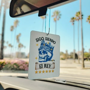 May include: White car air freshener with a blue skull wearing a crown and a banner that reads "SIGO SIENDO since '16 EL REY" with five gold stars below the banner.