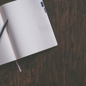 May include: An open notebook with lined pages and a black pen lying on a dark brown wooden surface.