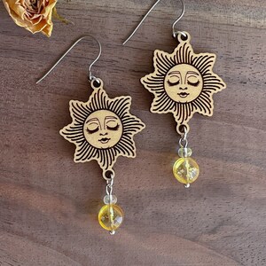 May include: A pair of wooden earrings with a sun face design and a yellow bead dangle. The earrings are made of light brown wood and have a silver hook.