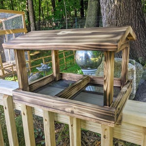 May include: A wooden bird feeder with a roof and two feeding stations. The feeder is stained a dark brown color and has a rustic look. It is mounted on a wooden railing.