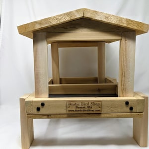 Puede incluir: Un comedero para p&aacute;jaros de madera con techo y plataforma. El comedero tiene una bandeja de metal negro y un letrero que dice "Rustic Bird Shop, Dracut, MA, www.RusticBirdShop.com".