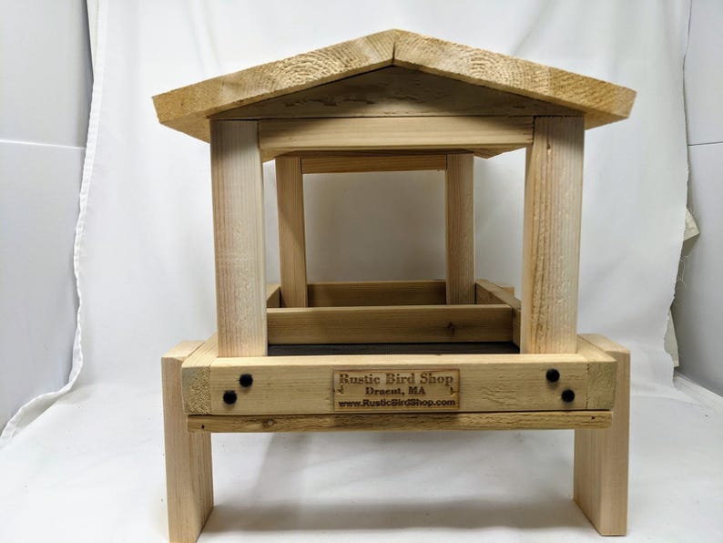 Puede incluir: Un comedero para p&aacute;jaros de madera con techo y plataforma. El comedero tiene una bandeja de metal negro y un letrero que dice "Rustic Bird Shop, Dracut, MA, www.RusticBirdShop.com".