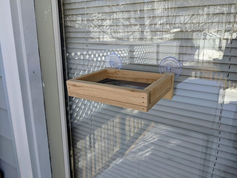 May include: A wooden bird feeder with a mesh bottom is attached to a window with two suction cups. The feeder is made of light brown wood and has a rectangular shape.