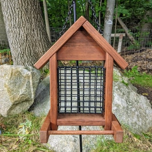 May include: A wooden bird feeder with a black wire mesh cage. The feeder is stained a dark brown color and has a small perch on the front. The feeder is hanging from a black chain.