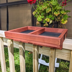 May include: A red wooden bird feeder with two compartments and a black mesh bottom. The feeder is mounted on a wooden railing.