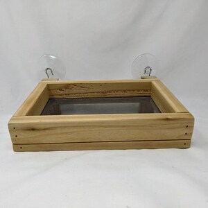 May include: A wooden bird feeder with a mesh bottom and two suction cups for attaching to a window. The feeder is made of natural wood and has a simple design.